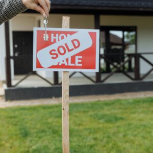 A hand holding a key in front of a sold sign and a house, symbolizing a successful real estate transaction.